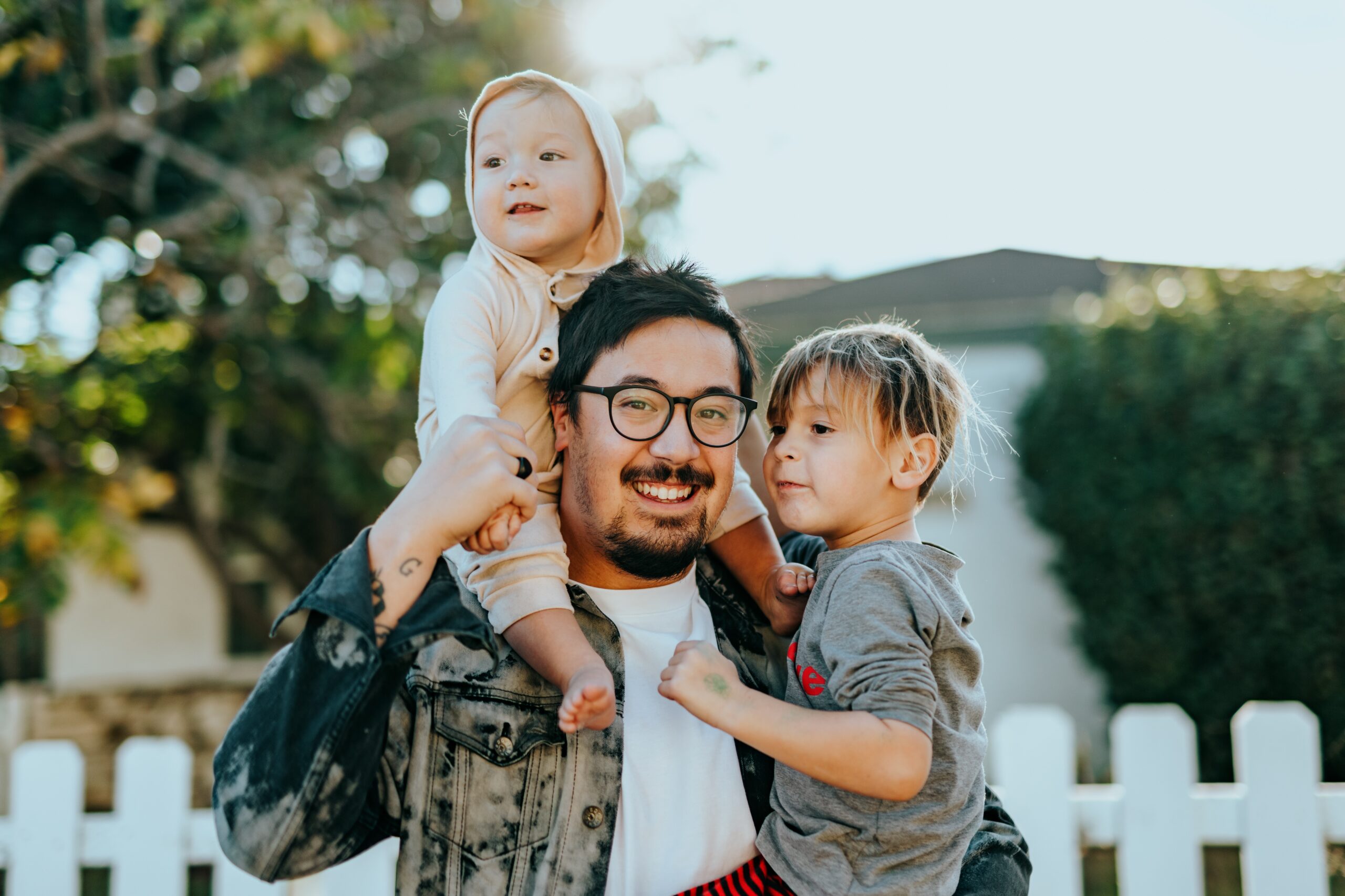 A father and 2 kids outside with a girl sitting on her father's shoulders.